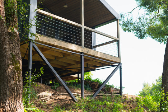 A modern cabin stands elevated on metal supports in a natural setting. The structure has a wooden deck and is framed by lush trees and vibrant greenery