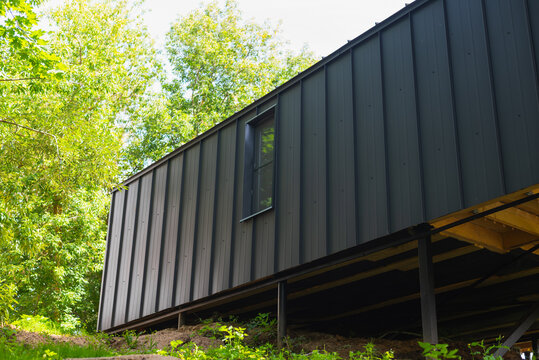 A black cabin is located on a hillside, elevated on stilts to protect it from ground moisture. Surrounding trees and greenery create a natural backdrop for this structure