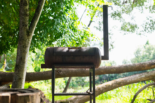 A metal grill is set on a stand in a lush outdoor area. Sunlight filters through the trees, creating a bright scene perfect for outdoor cooking