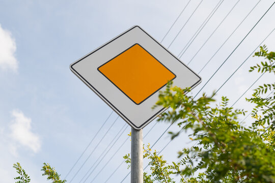 A road sign main road stands upright with a diamond shape and a yellow center against a blue sky