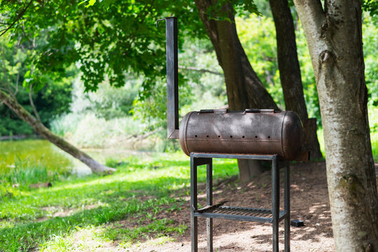 A metal grill stands on a sturdy frame under the shade of trees by a peaceful lake.