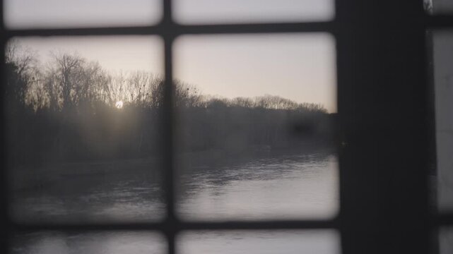 The sun rises low over a calm river, its glow diffused through an antique window and reflected softly on the water surface in Paris.