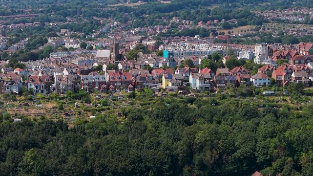 Belmont aerial approach toward Emmanuel church in Hastings, with surrounding hillside houses, showing dense residential rooftops, leafy woodland edge, and townscape depth in daylight.