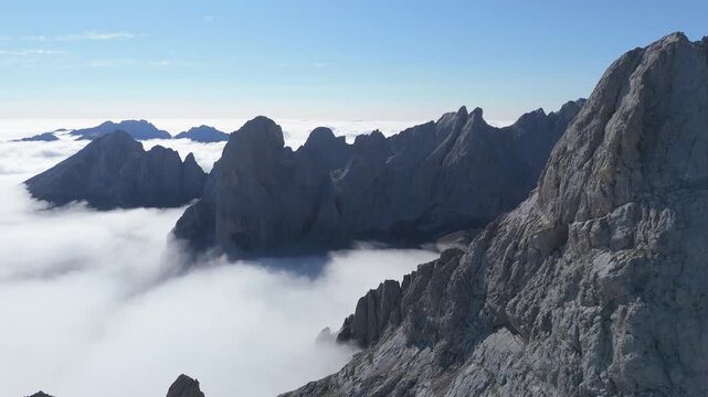 Aerial drone push in shot revealing the dramatic rocky peaks of Picu Urriellu mountain in Picos de Europa, Asturias, Spain, with a sea of clouds below and a clear blue sky above.