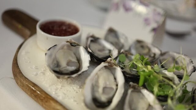 Appetizing close-up shot of fresh oysters on the half shell, artfully presented on crushed ice with vibrant microgreens and a tangy red dipping sauce, in soft natural lighting.