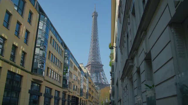 Eiffel tower Paris alley buildings blue sky backdrop, handheld shot. Scenic view of French capital landmark framed by traditional Parisian architecture.