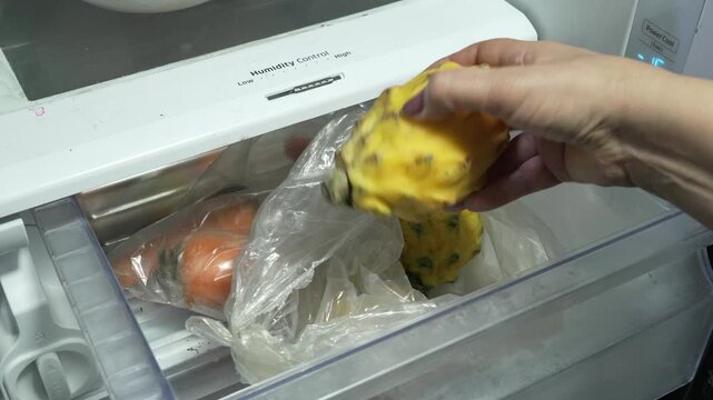 A person reaches into a fridge crisper drawer and pulls out a yellow dragon fruit from a plastic bag beside stored carrots. Useful for food storage, nutrition, or home lifestyle topics.