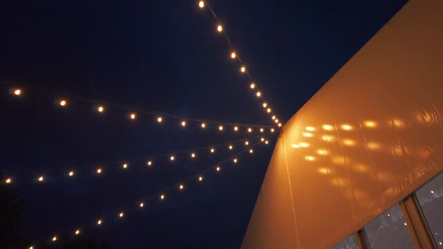 Warm string lights glowing against the night sky outside a modern event venue. Elegant outdoor decorative lighting creates a cozy atmosphere for weddings, parties, celebrations