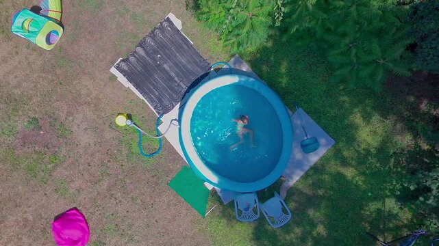 High angle top-down aerial shot captures a smooth spinning and descending drone movement over a pre-teen girl twirling in the clear blue water of a round inflatable pool in a sunny green garden.