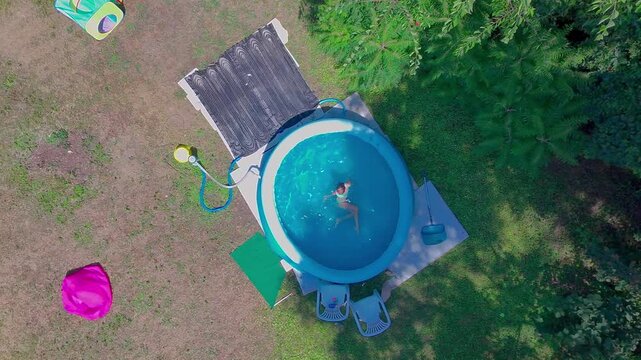 High angle top-down aerial shot captures a pre-teen girl floating and diving backwards under the clear blue water of a round inflatable swimming pool in a sunny green backyard garden.