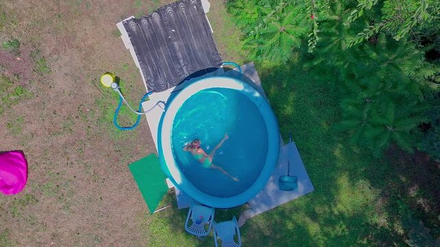 A high angle top-down aerial shot captures a pre-teen girl doing water exercises and swimming laps inside the clear blue water of a round inflatable pool in a sunny backyard garden during summer days.