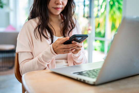 Asian female freelancer holding a mobile phone, messaging or checking notifications in front of a laptop