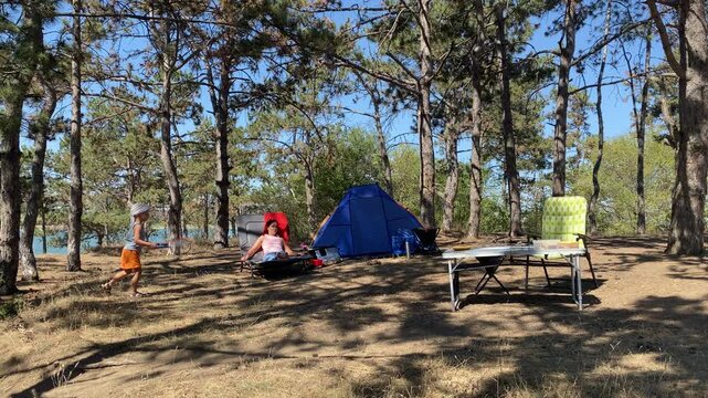 A young boy plays happily with a racquet while his mother rests on a chair. They enjoy a peaceful family campsite next to a blue tent in a pine forest. The sunny day feels warm and calm.