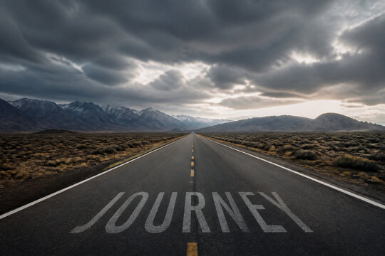 A long, empty road stretches toward distant mountains under a dramatic, cloudy sky.