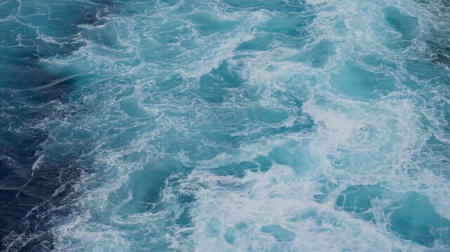 Aerial view of turbulent turquoise ocean water with white foam patterns swirling across the surface. Deep blue sea waves churn and mix creating mesmerizing natural textures and movement.