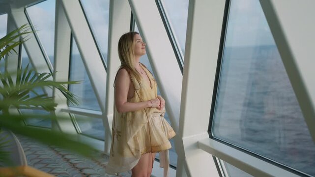 Young woman in flowing yellow summer dress gazes peacefully through geometric panoramic windows aboard luxury cruise ship, tropical palm fronds frame the scene as ocean horizon stretches beyond.