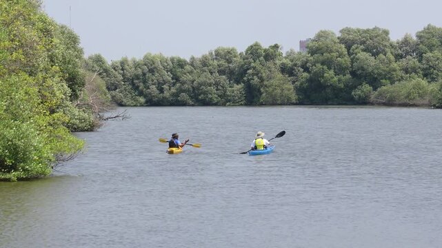 Two kayakers paddling yellow and blue single kayaks on a calm river framed by a dense green mangrove forest under a clear sky in Jakarta.