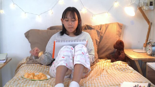 Relaxed young girl  sits comfortably on her bed, reading from a notebook while enjoying a healthy snack in a warm and cozy bedroom illuminated by soft decorative lights