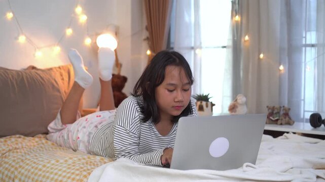 Young girl comfortably on bed using a laptop study online in bedroom illuminated by soft fairy lights	
