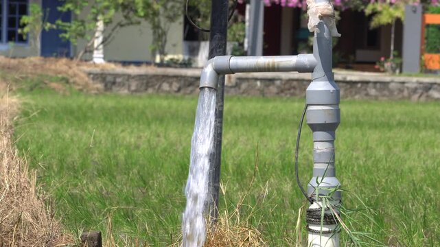 Water flows from a PVC pipe, pumping groundwater to irrigate a vibrant green rice paddy field, showing agricultural technology in a rural Southeast Asian farm landscape with a focus on splash.