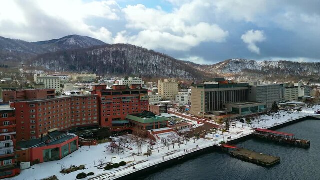 Smooth drone shot moving closer toward the snow-covered lakeside promenade, luxury resort buildings, and docks during winter season.