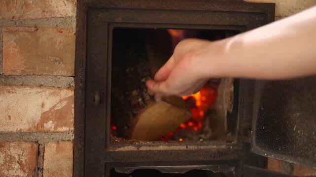 Close-up of a hand placing firewood into a traditional wood burning stove with glowing embers. Cozy rustic kitchen, heating and cooking with natural firewood.
