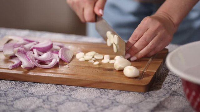 Close-up of hands slicing fresh garlic cloves and red onion on a wooden cutting board. Home cooking preparation with fresh ingredients for a rustic homemade meal.