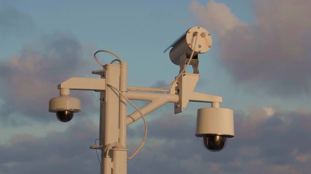 Surveillance cameras on cruise liner mounted on white metal pole with dual PTZ dome units and cables. Professional maritime security equipment against dramatic cloudy sky at dusk.