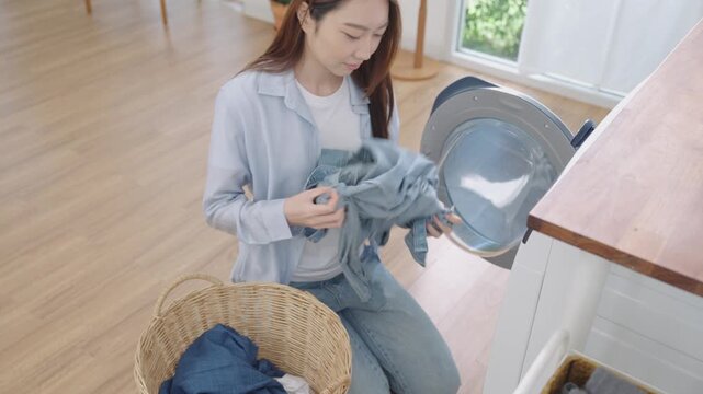 Young happy Asian Japanese woman unloading clothes, smell freshly washed shirt from washing machine or dryer, and put into basket in bright cozy laundry room at home in calm and peaceful morning