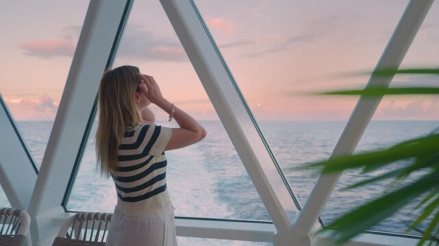 Young blonde woman in striped shirt stands on cruise liner deck gazing at pink sunset over Caribbean Sea through triangular glass windows with palm fronds and ocean wake visible.