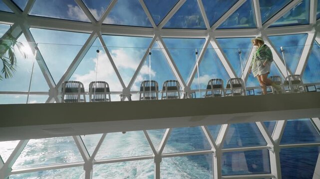 Woman in summer attire walks along a glass walkway on a modern cruise liner in Caribbean Sea. Geometric glass ceiling frames blue sky and turquoise ocean waves below creating stunning ship interior.