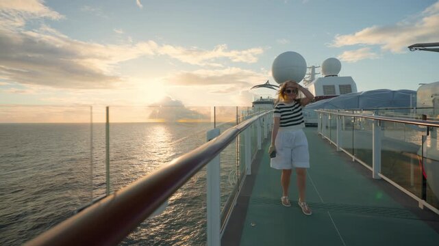 Woman in striped shirt walks along cruise liner deck in Caribbean Sea during stunning golden sunset. Glass railings and satellite domes frame the peaceful ocean voyage scene with warm light.