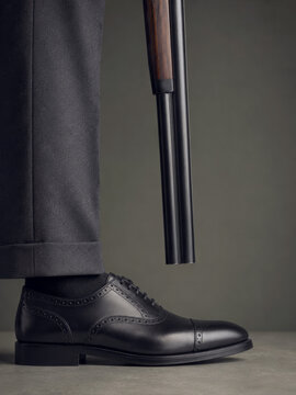 Humorous metaphor of undermining oneself with business man in black formal trousers and leather oxford shoe holding a barrel toward his foot
