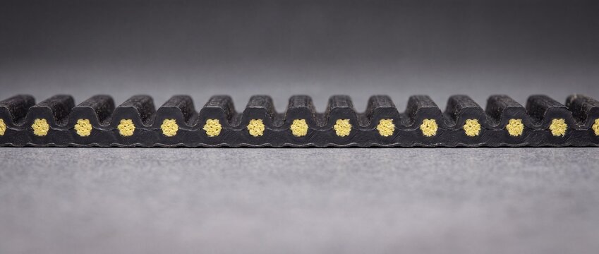 Cross section of reinforced timing belt showing yellow internal cords and black rubber teeth for automotive engine transmission.