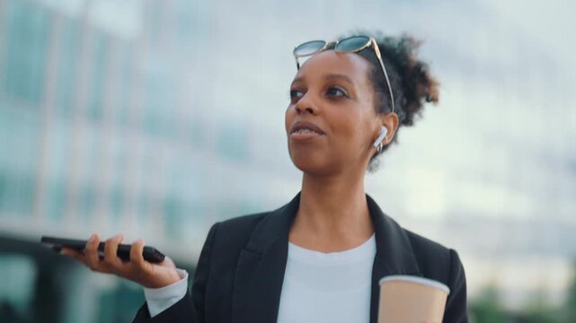 African american woman listening to voice message in phone, walking in city. Medium portrait of cheerful mixed race townswoman strolling alone in downtown, go to work in morning, working mobile call