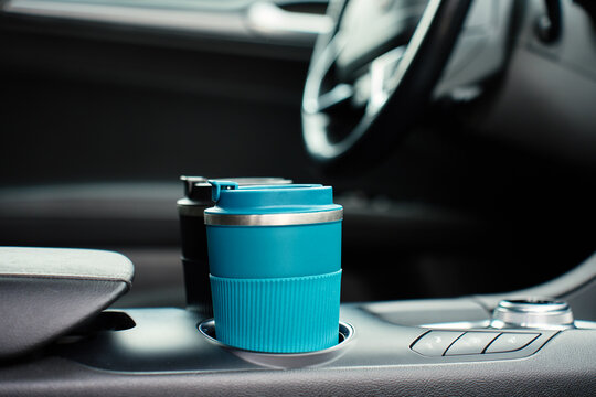 Two reusable coffee cups in the center console cup holders inside a modern car, with the steering wheel and dashboard blurred in the background