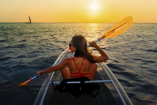 Silhouetted pretty sporty fit woman paddles clear kayak during vibrant Zanzibar sunset. Back view capturing peaceful golden hour on the Indian Ocean during a tropical tour with evening breeze
