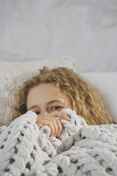 Cute woman with curly hair sits on bed with blanket pulled up to face. She smiles while peeking through fingers