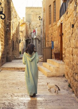 Woman in traditional abaya walking with cat through narrow stone alley in historic city of Mardin Turkey with ancient architecture