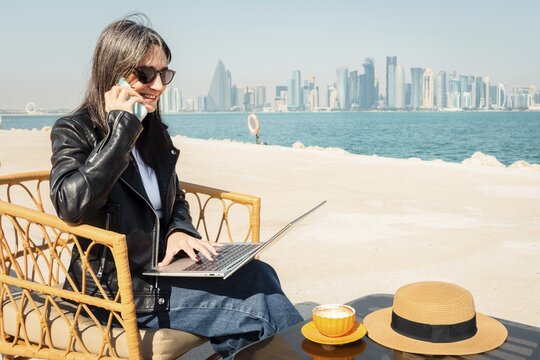 Happy business woman drinking coffee using laptop and smartphone chat. West Bay skyline background digital nomad working remotely in outdoor cafe. Middle East city urban professional