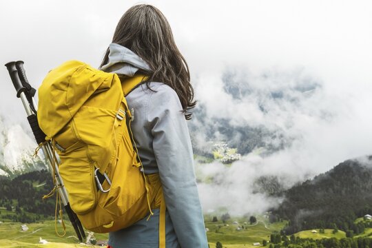 Back view of middle aged gray hair woman hiker yellow backpack overlooking lush green alpine valley misty mountains background Dolomites Alps Italy summer landscape