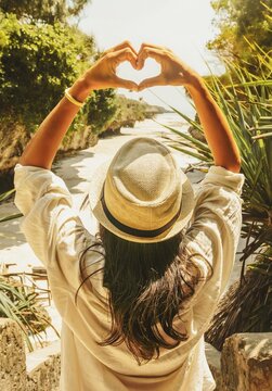 Young Woman tourist in straw hat framing heart sign with hands overhead looking down at tropical sandy cove beach surrounded by dramatic rocky cliffs vacation holiday lifestyle