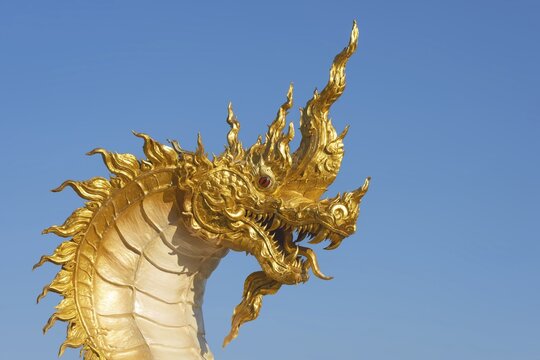 The golden Naga dragon rises against the clear sky of the Khao Kho Mountains. Its flame-like ornaments, red eyes and finely sculpted scales reflect the modern, mosaic-rich temple aesthetics for which this complex is famous. Buddhist temple complex Wat Phra That Sorn Kaew, Khao Kho Mountains, Phetchabun Province, Isan, Thailand