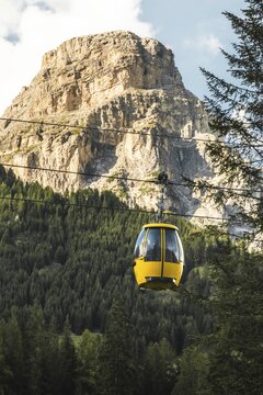 Dolomites, Italy - 27th july, 2026: Colfosco yellow cable car gondola lift ascending over lush green alpine meadows trail valley town pine forest towering mountain peaks Val Gardena Dolomites Italy