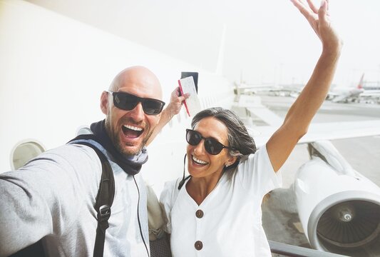 Cheerful excited caucasian couple taking selfie pose with boarding pass, passport in front of passenger airplane before boarding flight symbolizing travel, tourism, adventure, and vacation lifestyle
