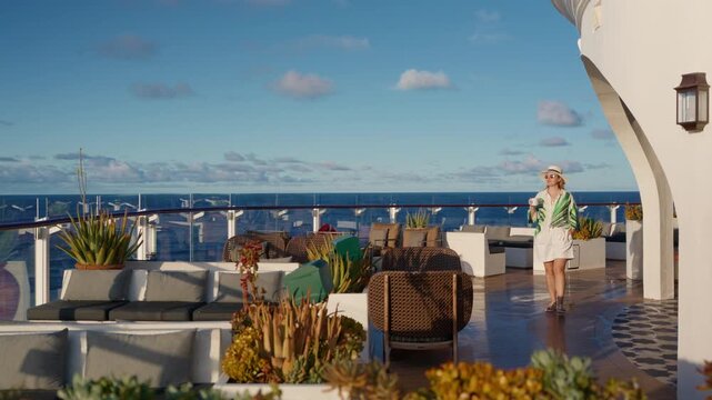 Stylish woman wearing straw hat and tropical palm print cover-up strolls across luxury cruise ship outdoor lounge deck with modern wicker furniture, succulent planters.