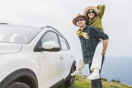Close up happy couple enjoys holiday time together piggyback ride by their white SUV during Mediterranean road trip. Lifestyle shot capturing love, freedom adventure on scenic coastal cliff side