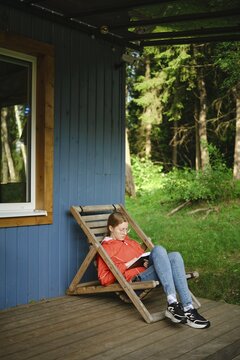 Young woman sits on wooden chair on terrace of tiny house and reading book. Trees and greenery surround her. It is daytime and the sun shines on the scene. She appears focused on her reading