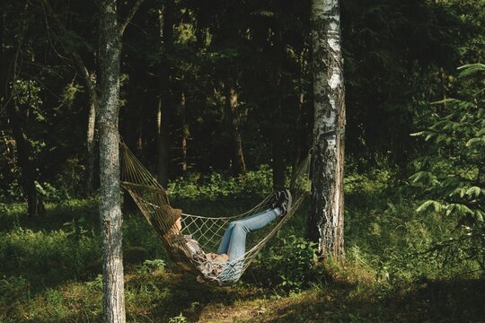 Young woman lies in hammock set up between two trees in woods. Sunlight filters through branches, creating shadows on ground. Surrounding greenery adds to natural setting