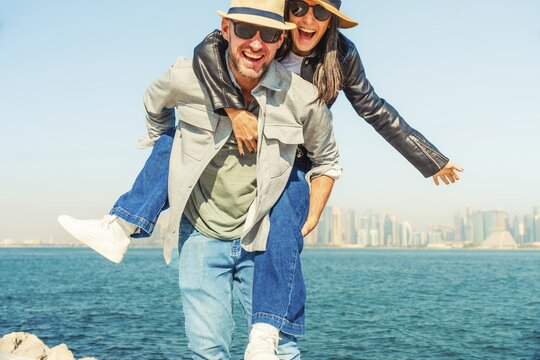 Happy tourist cute happy couple posing at bay. Man giving thumbs up, woman tongue out, sunglasses, hats, casual summer smart clothes. Skyline viewpoint by blue water on sunny day background